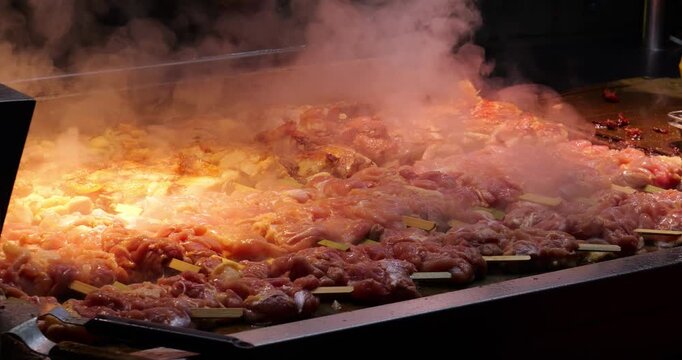 Medium shot of dozens of raw chicken meat skewers arranged closely together across huge heated flat-top griddle at Asian street food stall. Thick clouds of steam rise from searing poultry meat