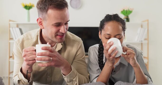 Couple drinking coffee talking on sofa together. Warm domestic scene of a diverse pair at home with tea, cozy light and calm mood, ideal for relationship blogs, lifestyle ads and social content. 