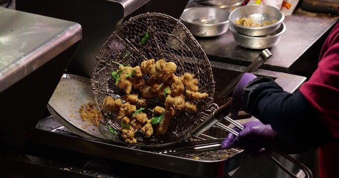 Taiwanese deep fried popcorn chicken being lifted out of deep fryer at night market food stall. Cook uses large wire mesh skimmer strainer basket to raise crispy breaded chicken chunks and greens