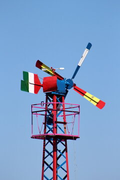Colorful vintage wind-powered mechanism once used to operate a water pump, with multicolored blades and an Italian flag tail standing against a clear blue sky.