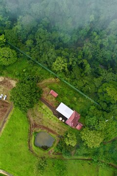 Aerial view of Kapok Hanging Bridges surrounded by lush rainforest, green fields, a small pond, and a building with a white roof in La Virgen, Heredia Province, Costa Rica.