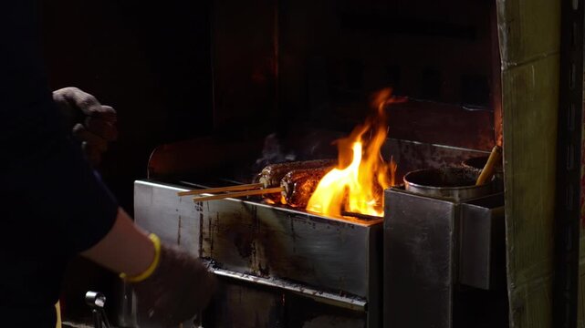 Close up shot of street vendor grilling roasted corn on stick over hot open fire at night market. gloved hands rotate corn cob as large orange flames burst upwards with smoke