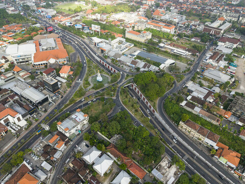 Aerial view of the Statue of Dewa Ruci at the busy roundabout intersection surrounded by urban buildings, roads, and lush greenery in Kecamatan Kuta, Bali, Indonesia.