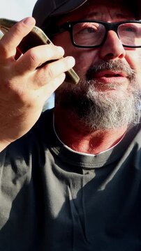A close-up of a middle-aged man with a beard and glasses actively engaged in a phone conversation, holding his smartphone to his ear in speakerphone mode, concluding with a warm and friendly smile.