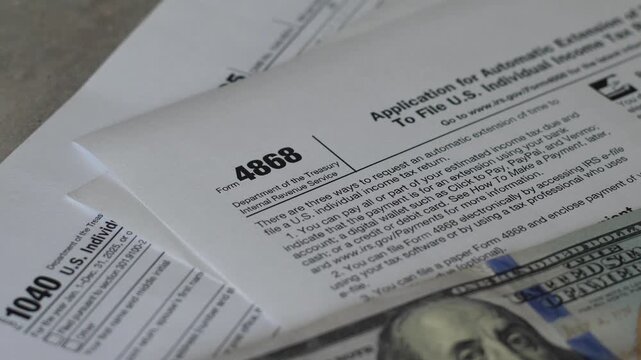 Close-up shot of US tax Form 1040 and Form 4868 with a stack of hundred-dollar bills placed on top. Official IRS paper documents prepared for financial filing,