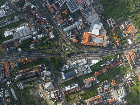 Aerial view of the Dewa Ruci Statue roundabout surrounded by dense urban buildings with orange rooftops and traffic flow in Kecamatan Kuta, Bali, Indonesia.