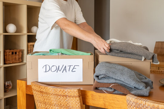 Person organizing and placing folded clothes into cardboard boxes with a donate label, preparing items for charity and decluttering home