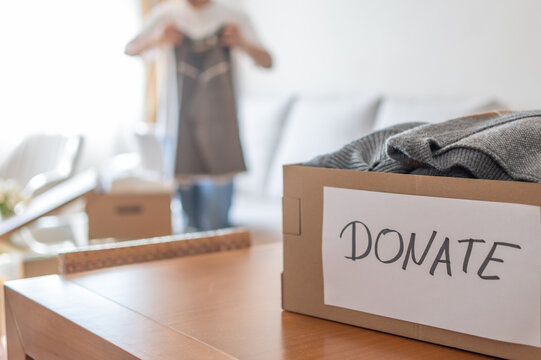 Person is decluttering home, collecting used garments into a cardboard box labeled donate to contribute to a charity drive, promoting sustainability and helping others