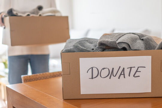 Person preparing cardboard boxes filled with pre-owned clothes, sweaters, and textiles, labeled donate, showing the concept of giving, decluttering, organizing, and helping others
