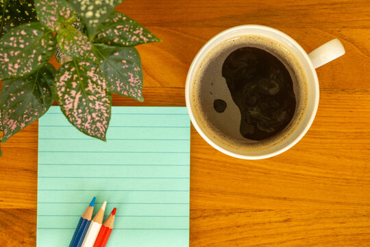 Vue de dessus d'une tasse de caf&eacute; noir, de crayons de couleur, de notes autocollantes vertes et d'une plante d&eacute;corative sur un bureau en bois, illustrant un espace de travail ou d'&eacute;tude chaleureux.
