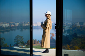 Fototapeta premium Female property manager standing on high rise terrace with coffee, showing scenic city and lake view. Concept of housing presentation, property development, urban living and business use