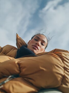 Woman in a warm tangerine orange puffer jacket and navy turtleneck, posing with a defiant gaze against a bright cyan sky for fashion campaigns