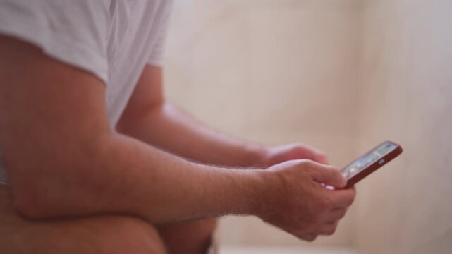 Side view of an unrecognizable man sitting on the toilet with his pants down, holding and scrolling through social media on his smartphone, highlighting phone addiction