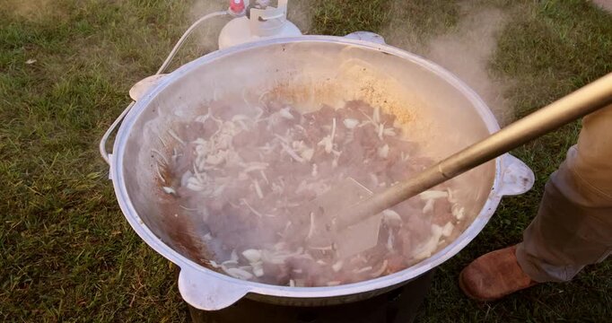 People cook meat in large pot at an outdoor gathering during chilly