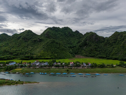 Aerial view of Phong Nha - near Ke Bang National Park with river, blue boats, houses, rice fields, and mountains under a cloudy sky. Phong Natural Heritage Area, Quang Tri, Vietnam.