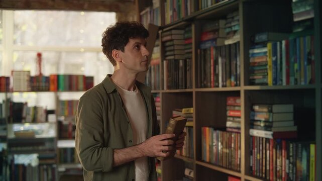 Young male scientist holds book while choosing another one from library shelf
