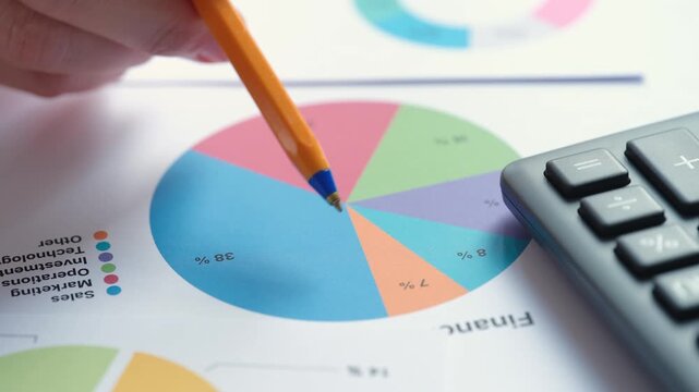 A hand analyzing a financial report by pointing an orange pen at a colorful pie chart next to a calculator. Close up.