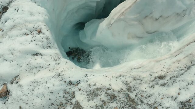 Aerial view of glacial meltwater carving through ice, revealing turquoise depths rocky debris. Cool tones dominate with white, blue, gray