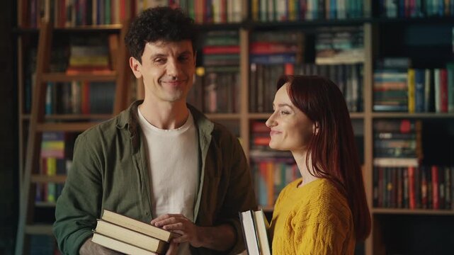 Positive red-haired woman and young man hold books and smile together in library