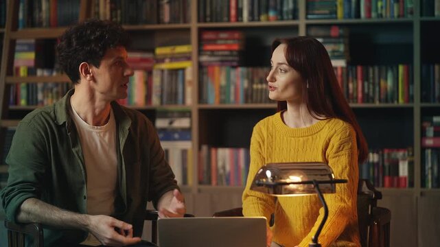 Male historian talks with colleague in library, conducting research on laptop