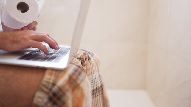Overworked businessman answering emails on laptop while sitting on toilet at home, illustrating poor work-life balance, excessive workload, stress and inability to disconnect from work.