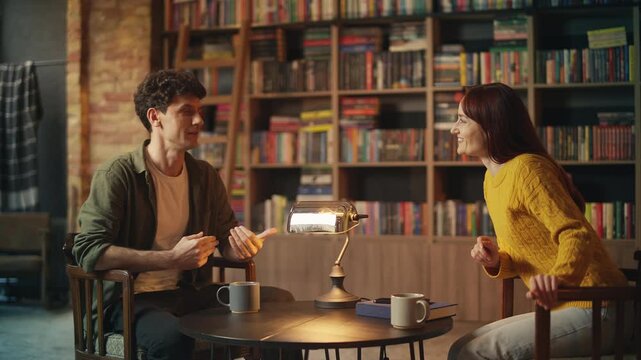 Positive young man and woman drink tea while chatting in cozy bookshop cafe