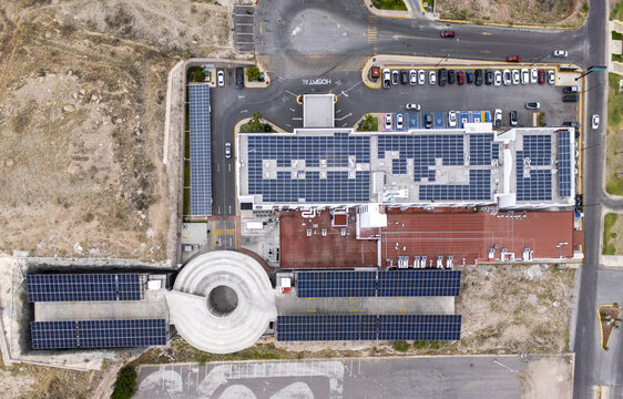 Aerial view of a hospital building featuring extensive rooftop solar panel arrays, a circular parking ramp, and a busy parking lot in Chihuahua, Chihuahua, Mexico.