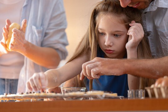 Focused young caucasian girl child baking sweet cookie dough at home kitchen table alongside father and mother parent family showing concentration learning domestic food cooking skill