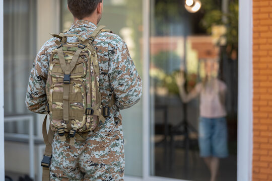 Brave soldier wearing camouflage uniform and tactical backpack stands outside glass door experiencing deep anticipation for an emotional family homecoming reunion