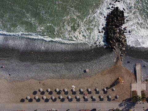 Aerial view of Costa Verde beach in Miraflores with rows of parasols on the pebble shore, crashing waves, and a stone breakwater, Miraflores, Provincia de Lima, Peru.
