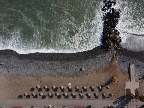Aerial view of Costa Verde coastline featuring a pebble beach, crashing ocean waves, a stone breakwater, and rows of beach umbrellas in Miraflores, Provincia de Lima, Peru.