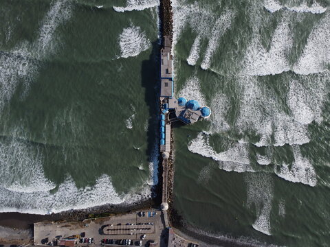 Aerial view of Restaurante La Rosa Nautica extending into the Pacific Ocean with crashing waves, blue roofs, and a parking area in Miraflores, Provincia de Lima, Peru.
