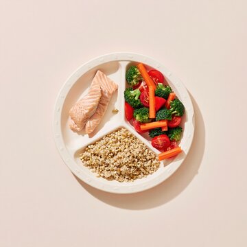 Divided portion plate with cooked salmon fillets, grains, and mixed vegetables on a pale beige background for dietetics and nutrition campaigns
