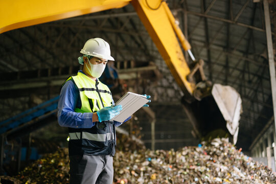 Industrial engineer with a digital tablet oversees waste sorting and recycling operations alongside heavy excavator machinery inside an urban disposal plant.