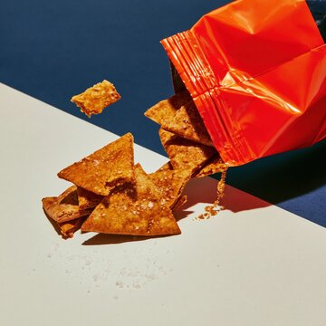 Red foil snack pouch spilling baked pita triangles on a white and blue background, featuring salty textures and crumbs for packaging mockup and product layout