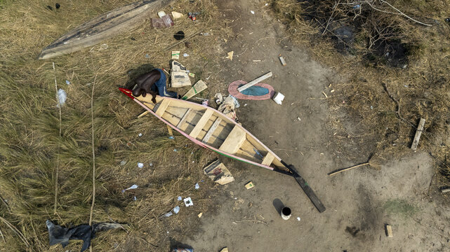 Aerial view of a man working on a wooden canoe near the shore of Sakumono Lagoon with scattered debris and dry grass in Tema Metropolitan, Greater Accra Region, Ghana.