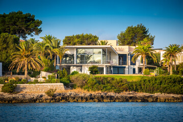 A modern luxurious seafront property located in Mallorca, Spain, with contemporary design, big glass windows. green garden with palm trees and the sea in front on a sunny day © lupiphoto
