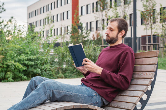 Young man reading book outdoors near modern residential building, combining education, self-development, leisure, mental wellbeing, work-life balance, student life, urban lifestyle