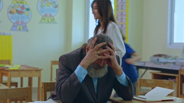 Classroom teacher student man with beard education school learning lesson adult male at desk showing stress worry and fatigue during document review in bright classroom setting