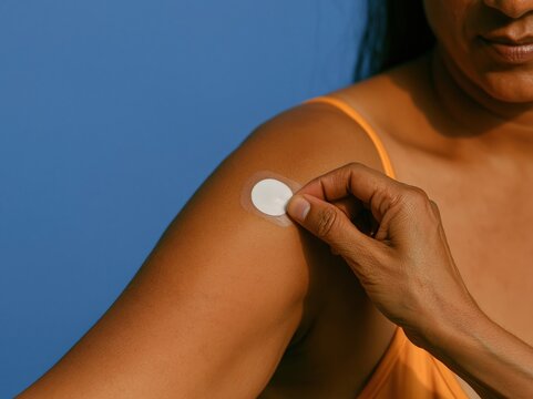 Hand of a South Asian woman applying a circular glucose sensor patch to her upper arm, featuring soft clinical lighting for medical mockup and health advertising