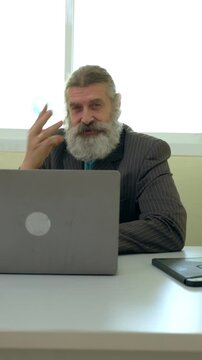 Senior man with beard using laptop at office desk in business suit, professional portrait with technology, meeting gesture near window bright workplace calm expression