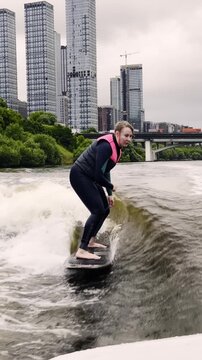 Person surfing on a wakeboard in a river near a city with tall buildings on a cloudy day