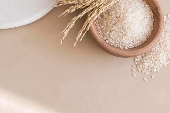 Wooden bowl of raw white rice grains with dried stalks on a warm brown surface, featuring soft textures and copy space for food packaging