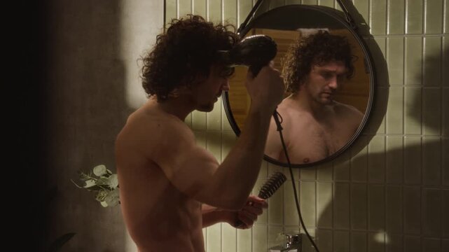 Young shirtless Caucasian man drying and brushing curly hair in front of bathroom mirror, seen from rear view. Footage is perfect for male grooming brands, hair care ads, and daily routine videos