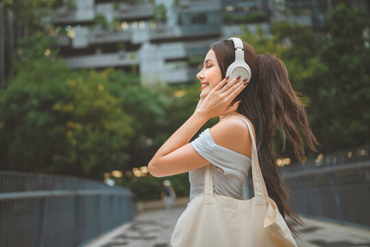 Happy young woman listening with headphones and carrying reusable tote bag outdoors, modern sustainable lifestyle
