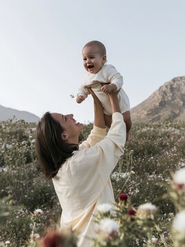 Mother lifting her laughing baby son high in a flowery meadow, featuring soft cotton textures and airy lighting for family and parenting concepts