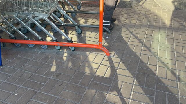 Worker's legs pushing a row of shopping carts on a paved sidewalk. Daily retail operations, logistics, or supermarket maintenance.