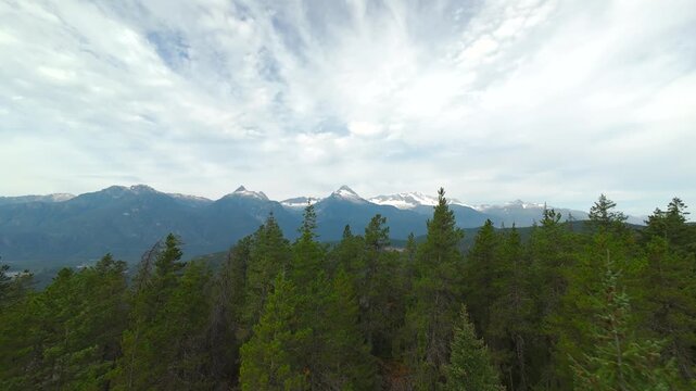 A wide aerial tracking shot glides over deep green treetops, showcasing the dramatic snow-capped peaks of Mount Tantalus under a bright sky with scattered clouds in the summer.