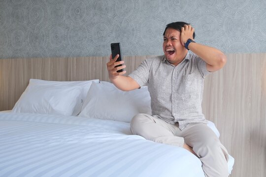 Shocked Asian man sitting on a bed, holding a smartphone and touching his head while reacting with surprise to something on the screen.
