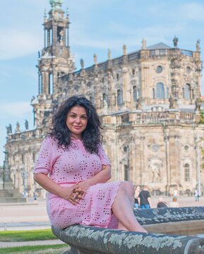 Beautiful brunette woman in pink dress sitting near fountain in Dresden, enjoying summer vacation with historic cathedral background in Saxony, Germany 